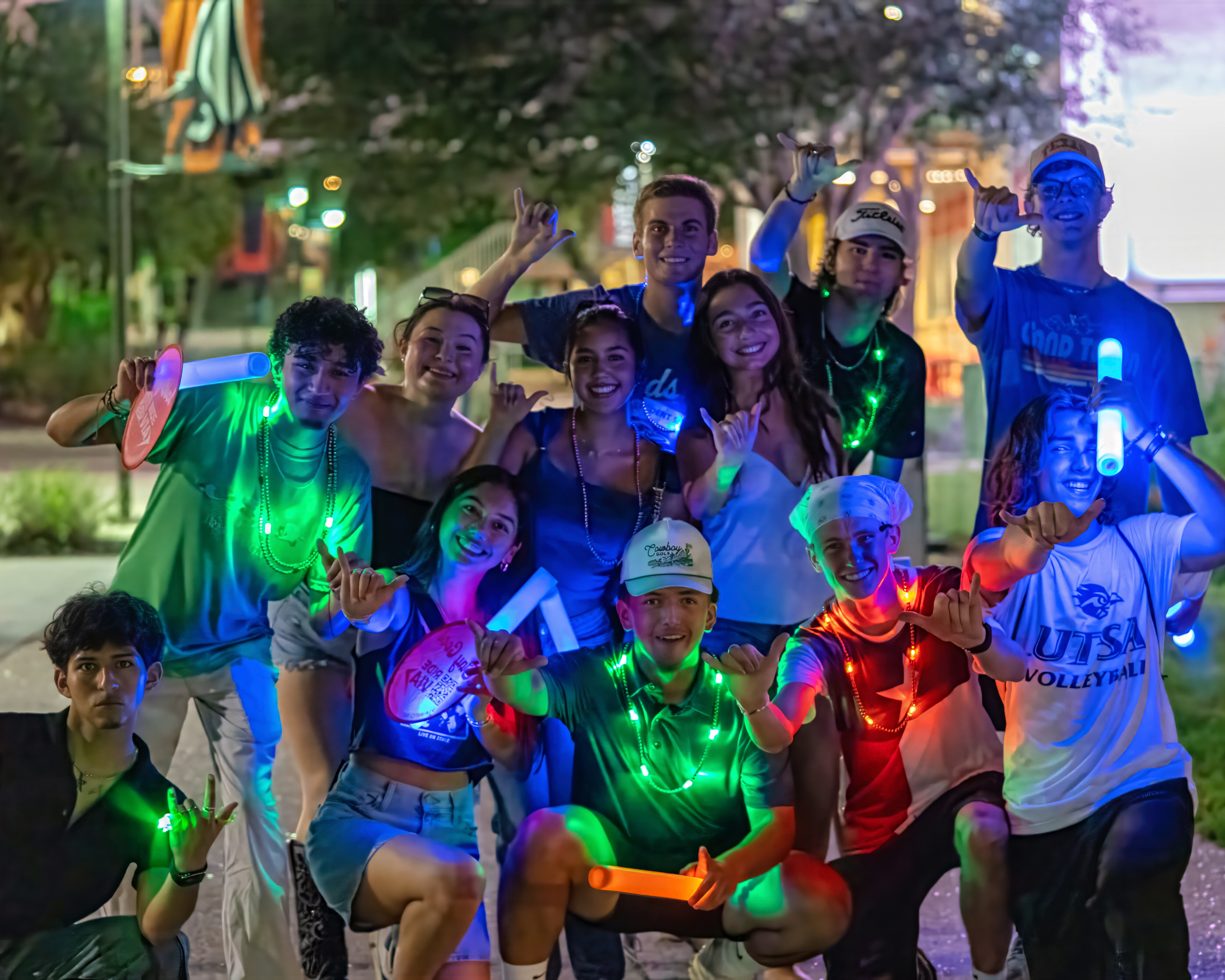 Multiple students with glowsticks and light up accessories posing for a photo with the UTSA birds up hand gesture.
